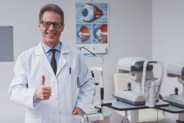 Handsome middle aged ophthalmologist showing OK sign and smiling while standing in his office - Fotoğraf, Görsel