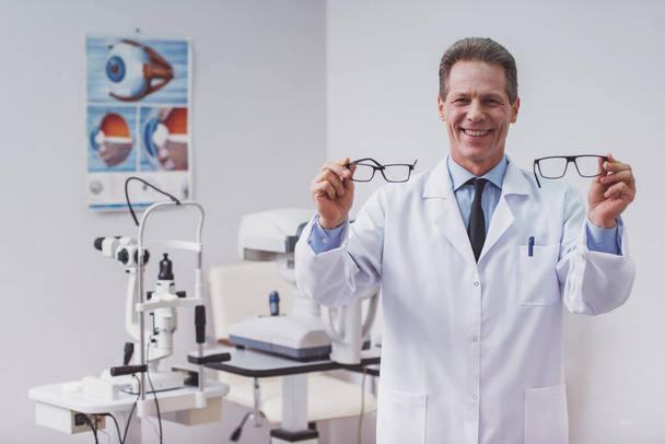 Handsome middle aged ophthalmologist holding eyeglasses and smiling while standing in his office - Foto, imagen