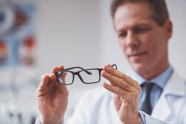 Bonito oftalmologista de meia idade segurando óculos e examinando-os, foco em óculos, close-up
 - Foto, Imagem