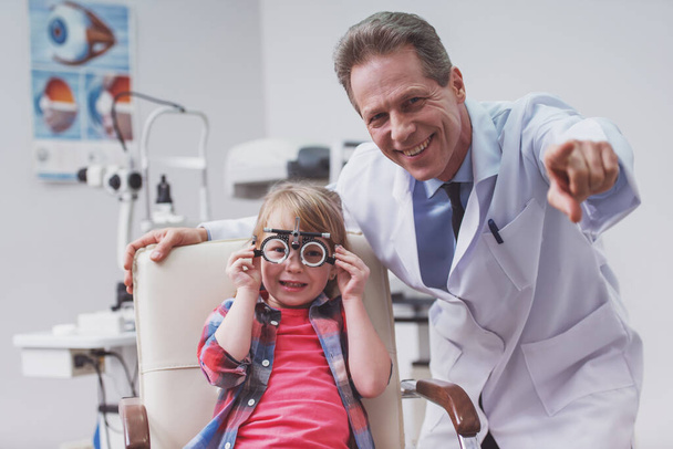 Красивый офтальмолог средних лет осматривает маленького мальчика с современным оборудованием, оба улыбаются
 - Фото, изображение