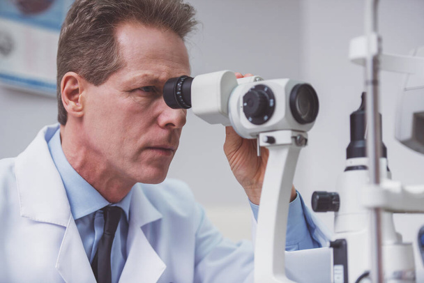 Portrait of handsome middle aged ophthalmologist examining patient with modern equipment - 写真・画像