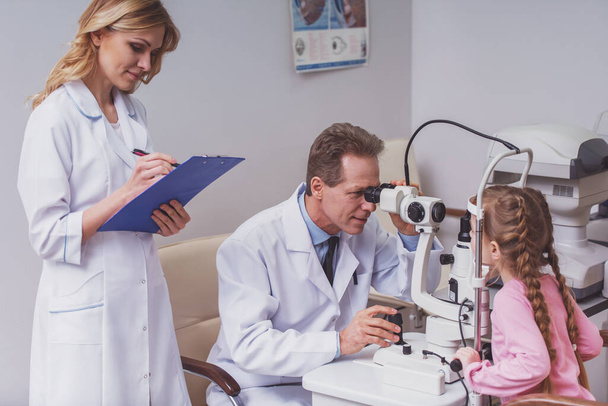 Handsome ophthalmologist examining little girl's eyes with modern equipment. Beautiful nurse making notes - Photo, image