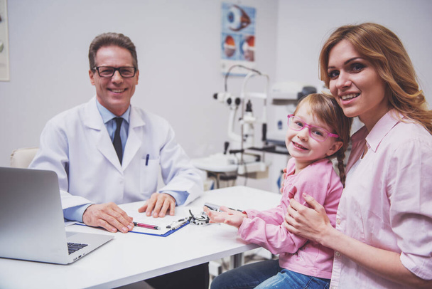 Bella bambina e sua madre all'oftalmologo su consultazione. Tutti guardando la macchina fotografica e sorridendo
 - Foto, immagini