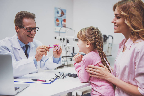 Nice little girl and her mother at the ophthalmologist on consultation, doctor holding glasses, all smiling - Zdjęcie, obraz