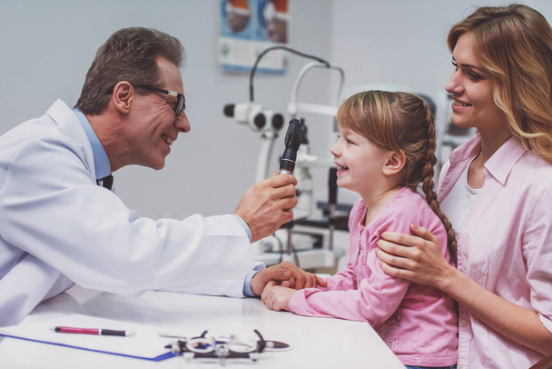 Menina agradável e sua mãe no oftalmologista, médico examinando menina com equipamento moderno, todos sorrindo
 - Foto, Imagem