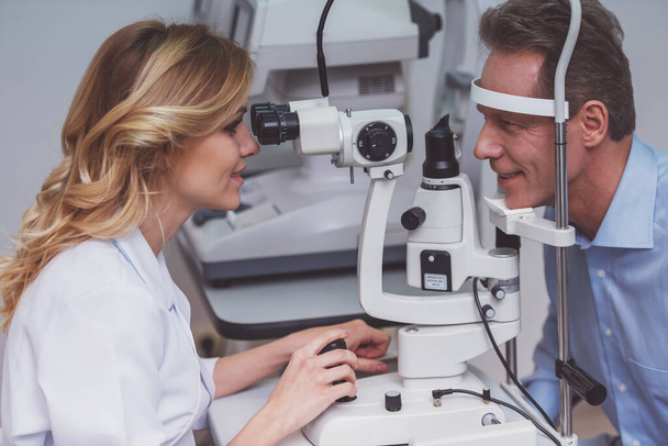 Young beautiful female ophthalmologist examining handsome middle aged man with modern equipment, both smiling - Photo, Image