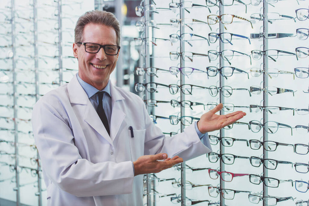 Knappe middelste leeftijd oogarts aanbieden van brillen, camera kijken en glimlachen terwijl staande in zijn kantoor - Foto, afbeelding