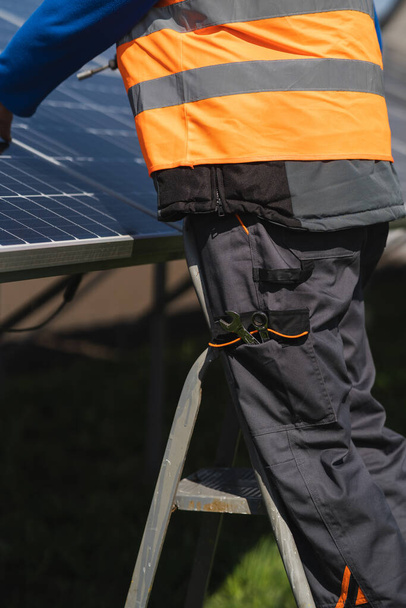 Férfi mérnök, aki napelemeket telepít és tart karban, egy létrán áll, és kivesz egy csavarkulcsot a zsebéből. - Fotó, kép