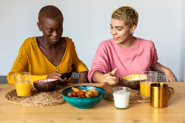 Happy multiethnic female couple relaxing at home while having breakfast - Fotó, kép
