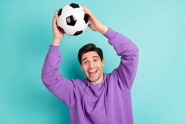 Photo of lucky funny young man wear purple hoodie smiling catching football ball isolated teal color background - Photo, Image