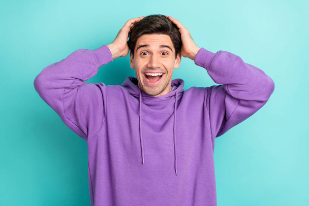 Photo of shocked funky young guy dressed violet sweatshirt arms head open mouth smiling isolated turquoise color background - Photo, Image