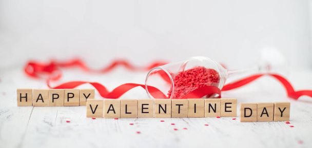 Happy St. Valentine Day greetings card. Wooden letters on the white background with small scattered red hearts and overturned champagne glasses - 写真・画像
