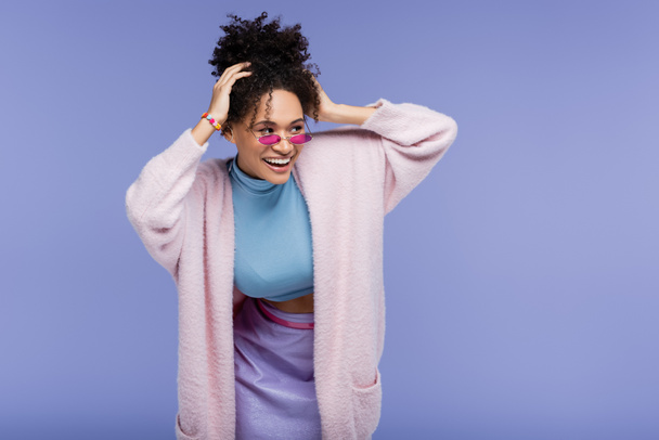 весёлая африканская американка в стильных солнцезащитных очках, регулирующая вьющиеся волосы, изолированные на фиолетовой  - Фото, изображение