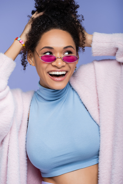 cheerful african american woman in pink sunglasses adjusting curly hair isolated on violet  - 写真・画像