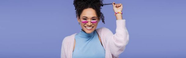 joyful african american woman in pink sunglasses pulling curly hair isolated on violet, banner - Photo, Image