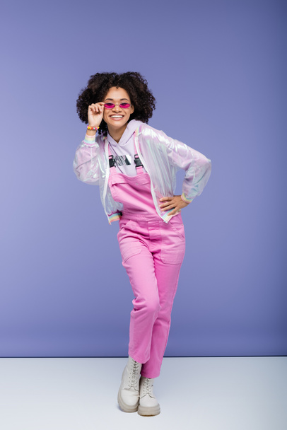 happy african american woman in pink overalls adjusting stylish sunglasses while posing with hand on hip on purple  - Foto, afbeelding