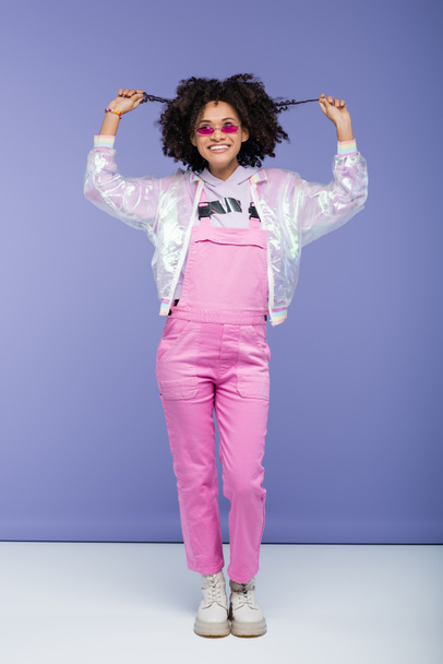 full length of happy african american woman in pink sunglasses and overalls pulling curly hair on purple - Φωτογραφία, εικόνα