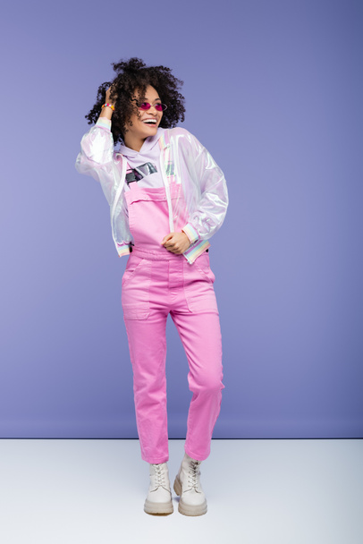 full length of smiling african american woman in sunglasses and overalls adjusting curly hair on purple - Foto, imagen