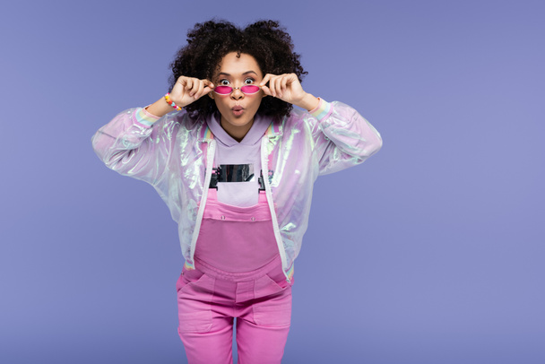 surprised african american young woman in pink overalls adjusting sunglasses isolated on purple - Φωτογραφία, εικόνα