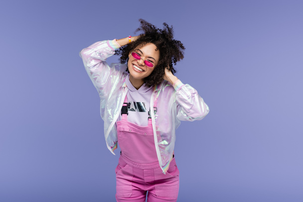 positive african american woman in stylish sunglasses and overalls adjusting curly hair isolated on purple - Fotografie, Obrázek