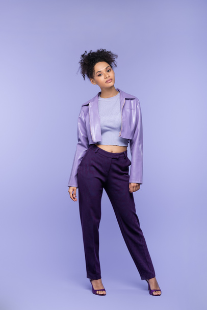 full length of curly and stylish african american woman in violet leather jacket and trousers posing on purple - Фото, изображение