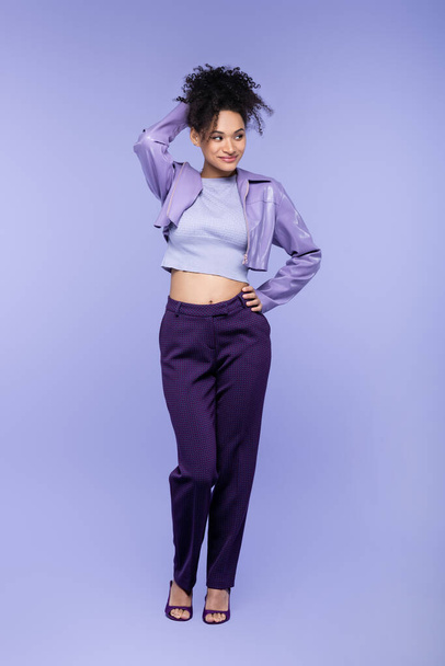 full length of flirty african american woman in violet leather jacket and trousers posing with hand on hip on purple - Foto, Imagem