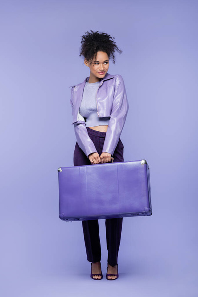 full length of smiling young african american woman holding suitcase on purple  - Photo, Image