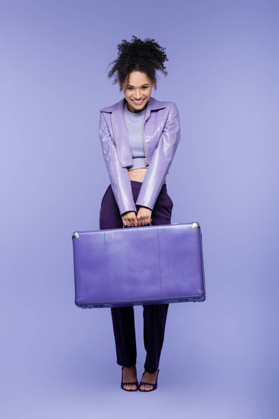 full length of cheerful african american woman holding luggage on purple  - 写真・画像