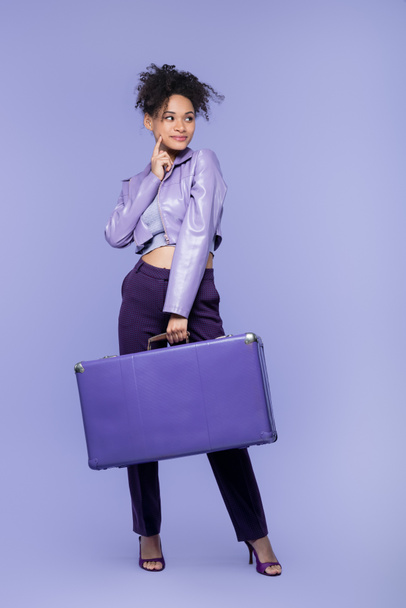 volledige lengte van pensive jong Afrikaans amerikaanse vrouw met bagage op paars  - Foto, afbeelding