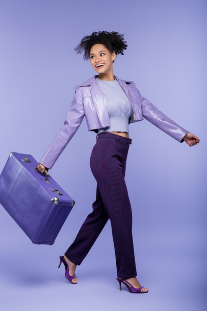 longitud completa de la mujer afroamericana feliz caminando con la maleta en púrpura  - Foto, Imagen