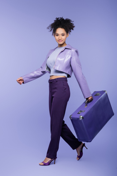 longitud completa de rizado joven afroamericana mujer caminando con maleta en púrpura  - Foto, Imagen