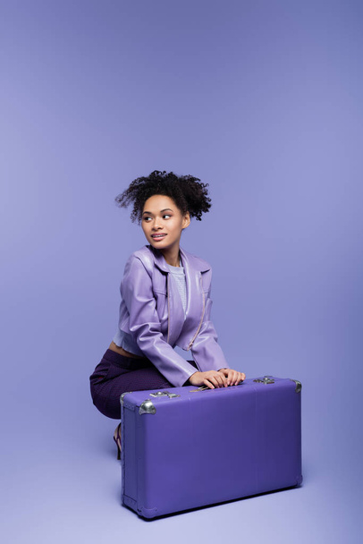 full length of curly young african american woman sitting near baggage on purple  - Фото, изображение
