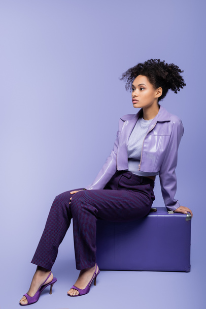 full length of young african american woman sitting on suitcase on purple  - Foto, afbeelding