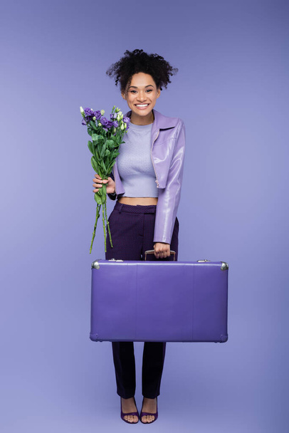 full length of smiling african american woman holding baggage and bouquet of flowers on purple  - Valokuva, kuva