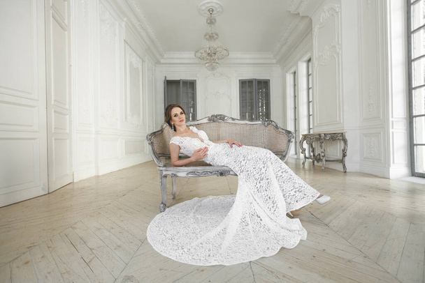 Beautiful bride posing in wedding dress in a white photo Studio. Mirror. Sofa. Bouquet. The door. Chandelier. - 写真・画像
