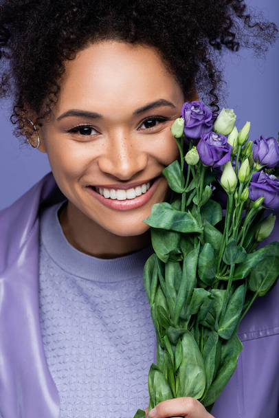 positive and curly african american woman near bouquet of violet flowers isolated on purple - Photo, Image