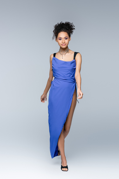 piena lunghezza di elegante donna afroamericana in abito blu brillante che cammina sul grigio - Foto, immagini