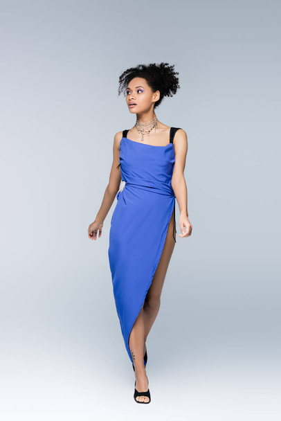 full length of curly african american woman in bright blue dress walking on grey - Zdjęcie, obraz