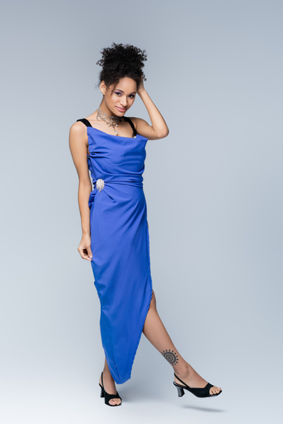 full length of smiling and tattooed african american woman in bright blue dress on grey - Photo, Image