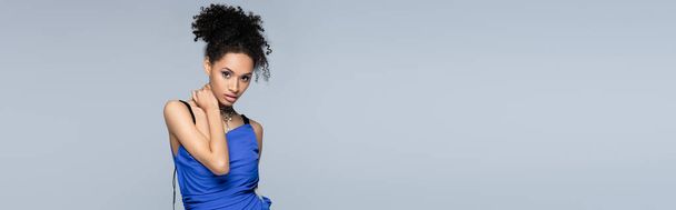 curly african american model in bright blue dress looking at camera while posing isolated on grey, banner - Foto, Bild