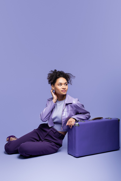 full length of dreamy african american model in leather jacket sitting near violet suitcase on purple  - Фото, изображение