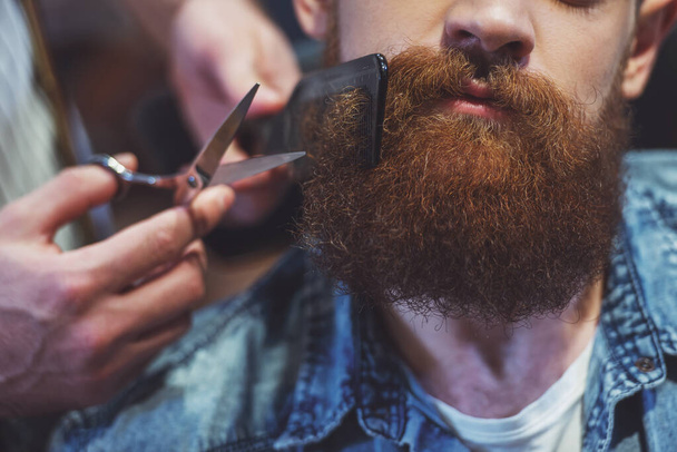 Attractive muscled bearded barber cutting handsome young customer beard at the barber shop, close-up - Photo, Image
