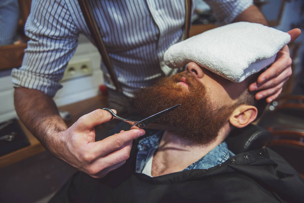 Привлекательный мускулистый бородатый парикмахер стрижет красивую молодую клиентскую бороду в парикмахерской
 - Фото, изображение