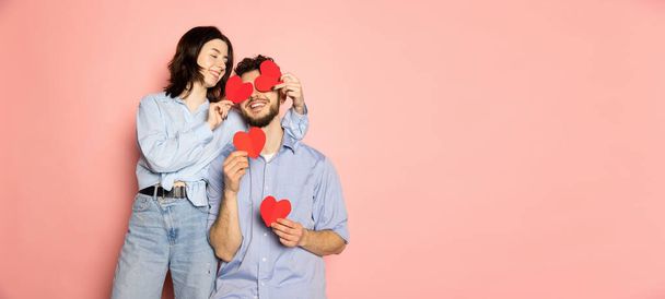 Flyer. Young and happy man and woman holding greeting cards shaped hearts isolated on pink trendy color background. Emotions, youth, love and lifestyle concept - Фото, зображення
