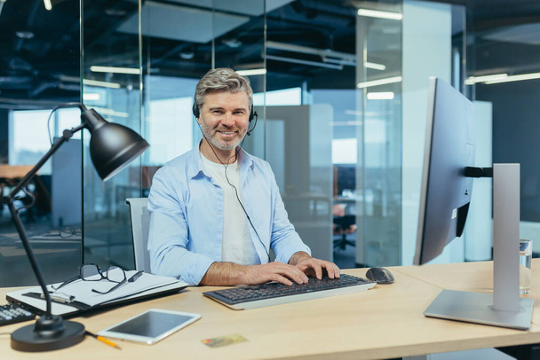 Retrato de un exitoso hombre de negocios, canoso hablando con colegas en una videollamada, usando un auricular, sonriendo y regocijándose - Foto, Imagen