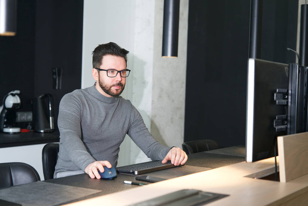 Portrait of serious concentrated bearded guy, young focused man freelancer in glasses is working from home, apartment on pc computer in glasses, looking at monitor. Freelance distant job, worker - Foto, Bild
