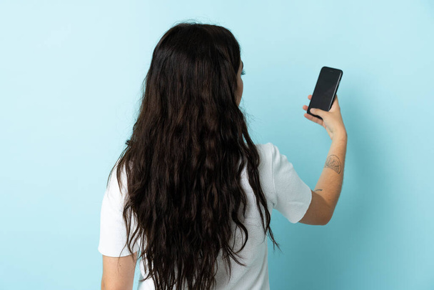 Молода жінка використовує мобільний телефон ізольовано на синьому фоні в задньому положенні
 - Фото, зображення