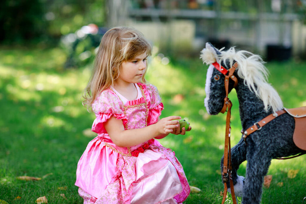 Little preschool girl hugging with rocking horse toy. Happy child in princess dress on sunny summer day in garden. Girl in love with her favourite old vintage toy animal. - Photo, Image