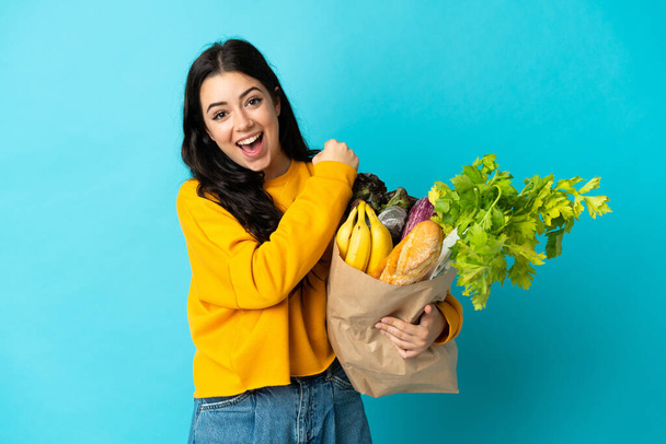Fiatal nő kezében egy élelmiszer bevásárló táska elszigetelt kék háttér ünnepli a győzelem - Fotó, kép