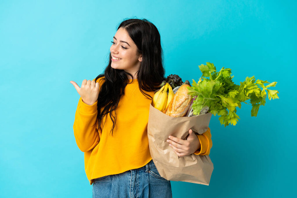 Elinde alışveriş torbası tutan genç bir kadın mavi arka planda izole edilmiş bir şekilde bir ürünü sunmak için yan tarafı işaret ediyor. - Fotoğraf, Görsel